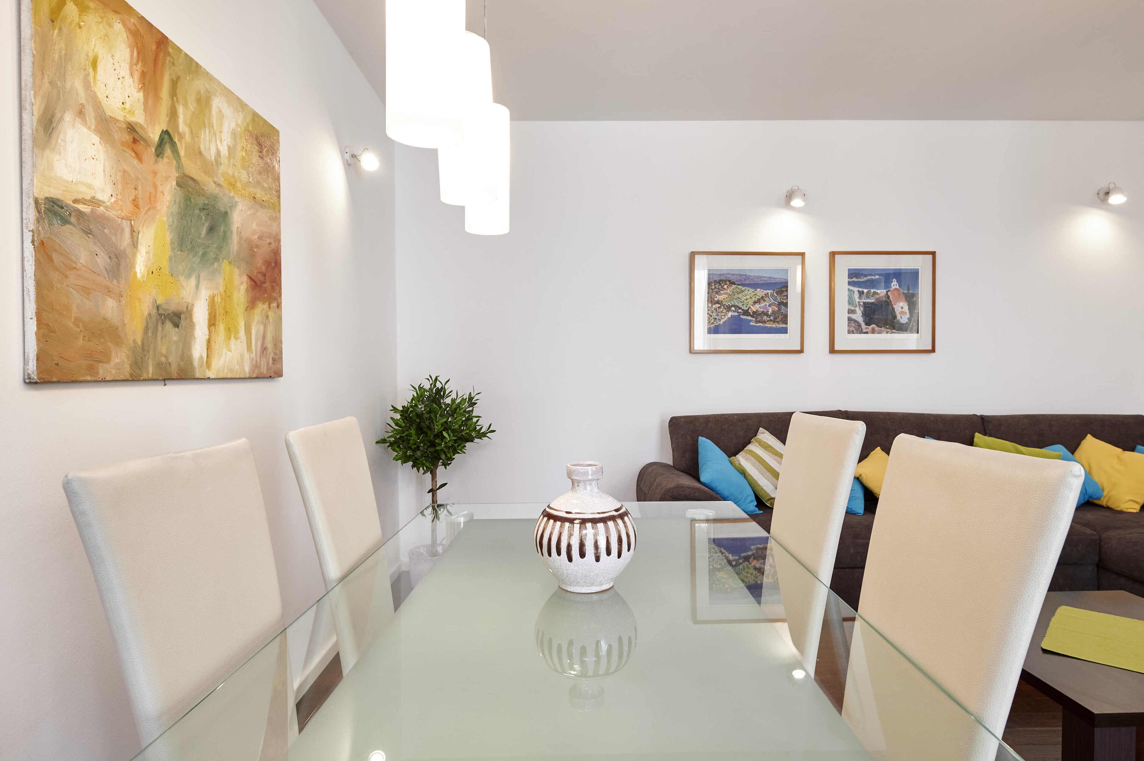 A modern living room with a glass table, white chairs, a brown sofa with colorful cushions, and two framed pictures on the wall.