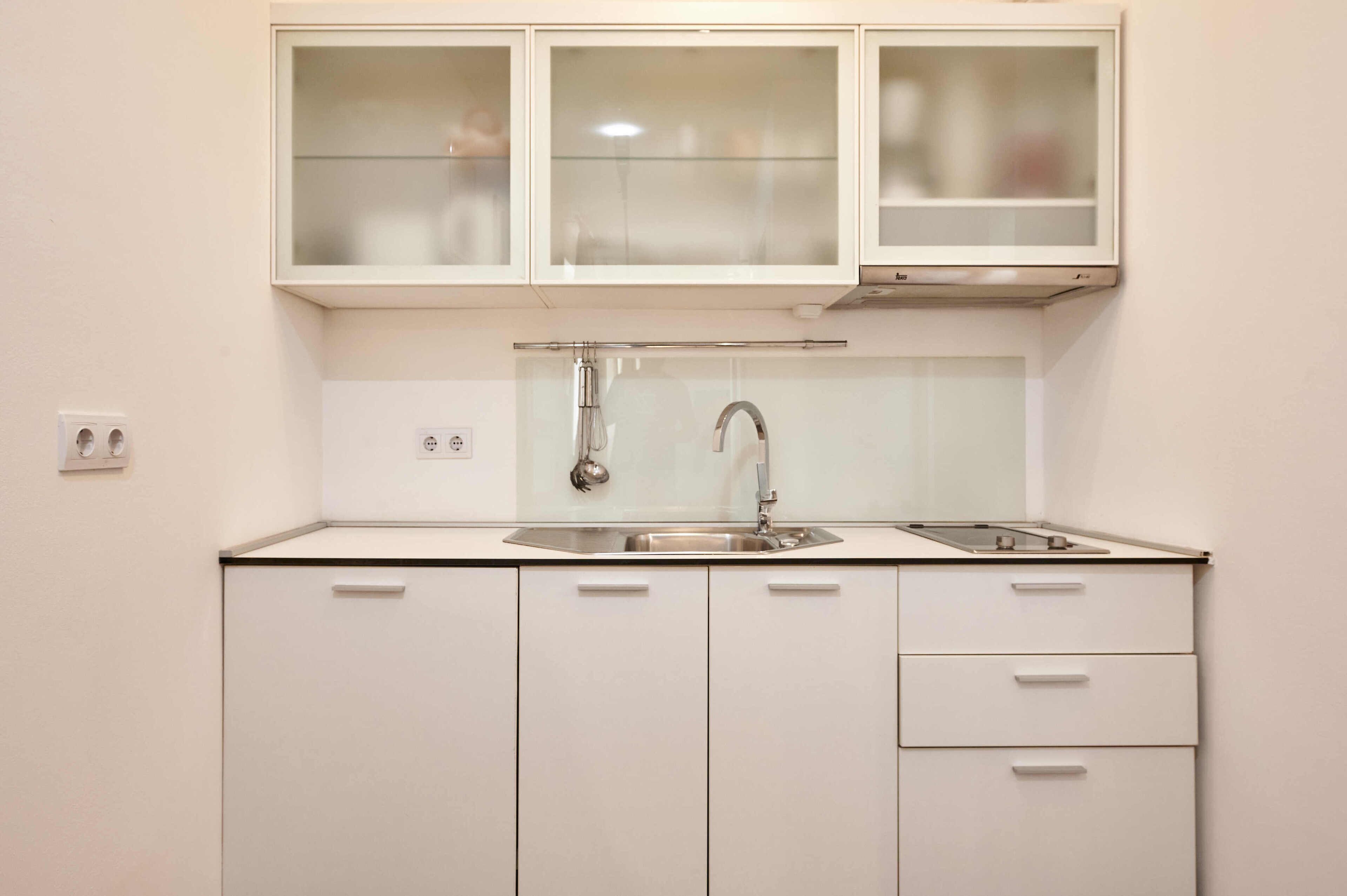 Cocina compacta con pileta de acero inoxidable, grifería doble y horno integrado. Se observan tres armarios con puertas de vidrio y un cajón.