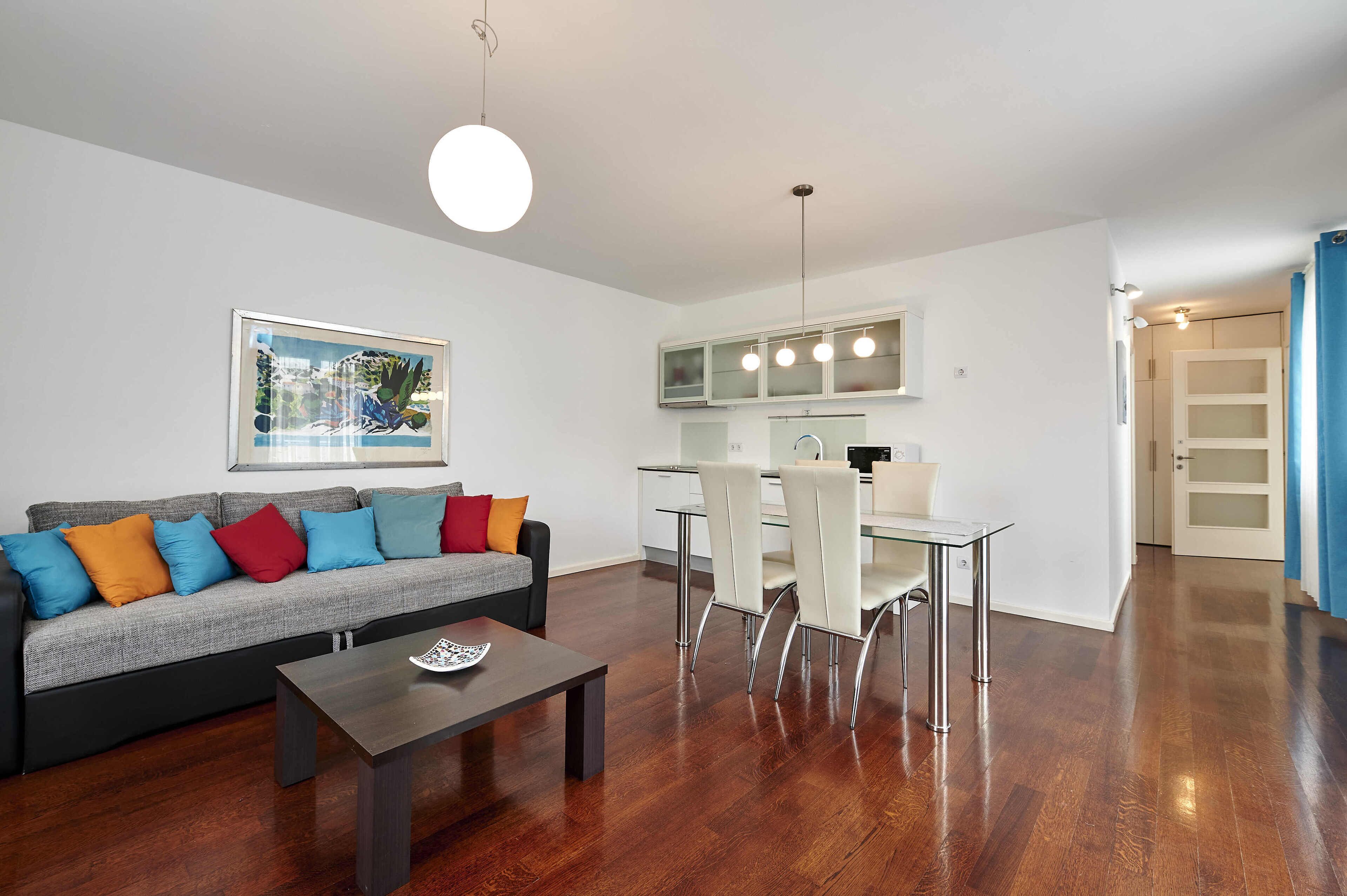 A modern living room with a grey sofa, colorful cushions, a wooden coffee table, and a dining area with a glass table and white chairs.