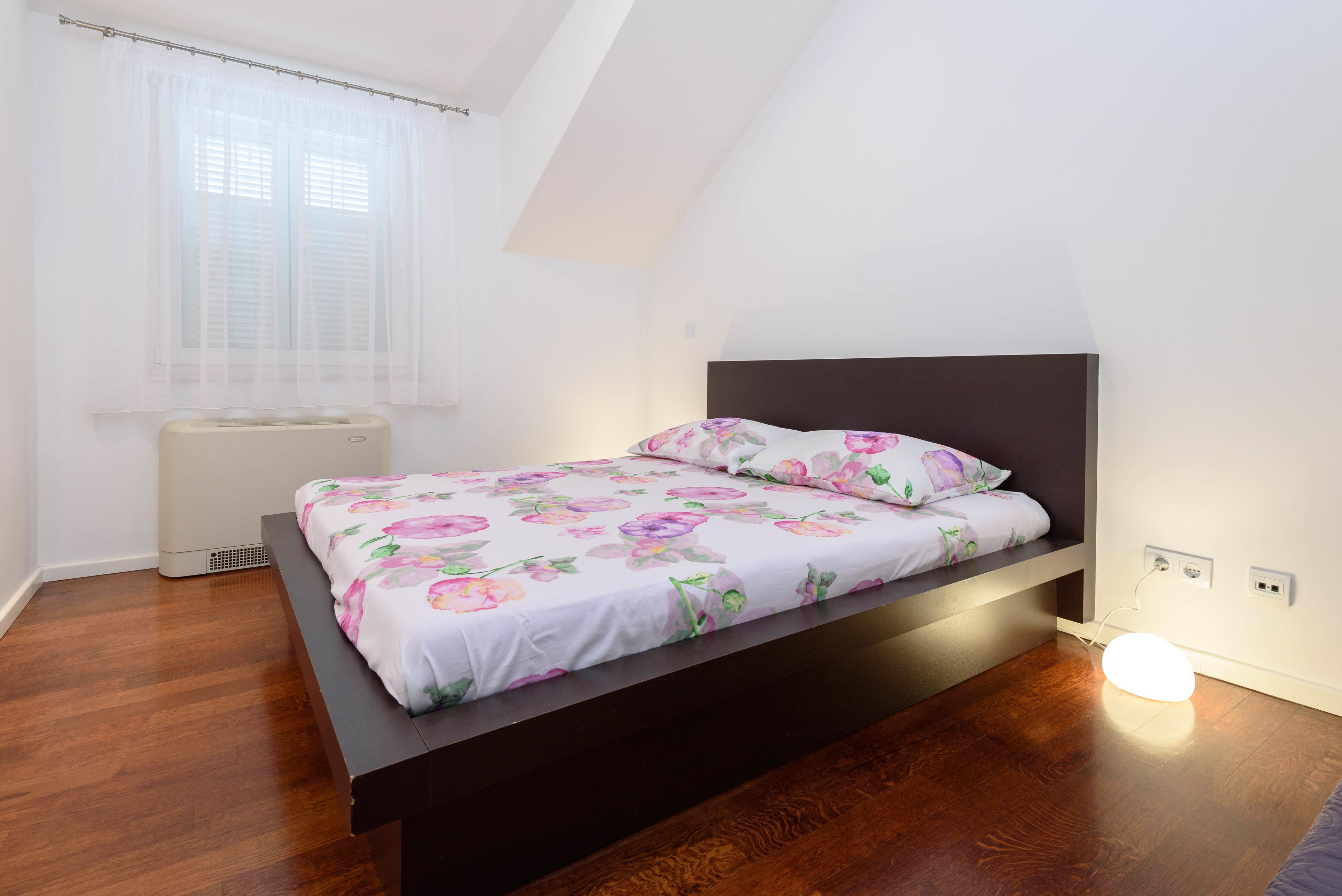 A bedroom with a bed, a window with curtains, a radiator, and a wall socket.