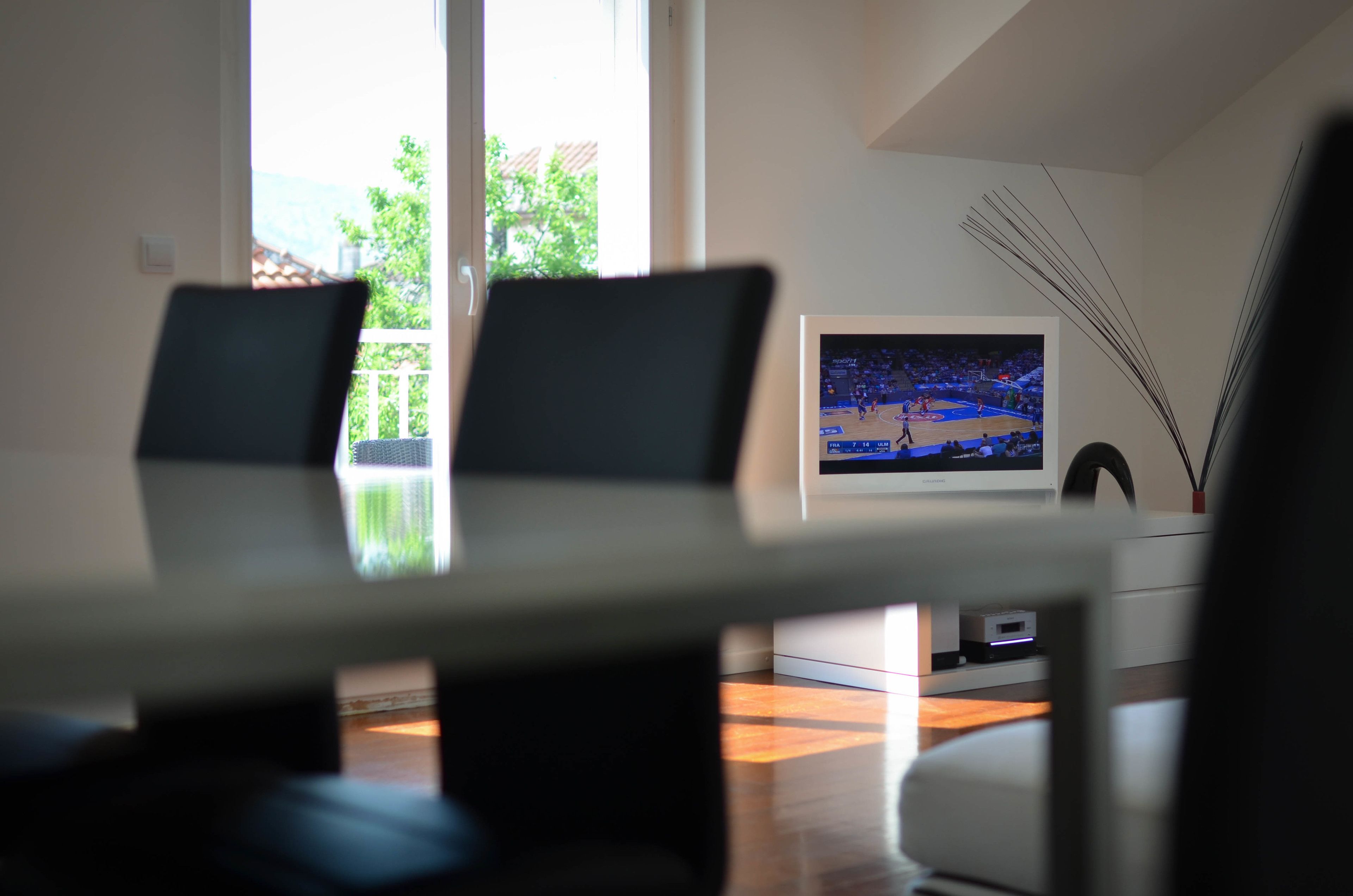 A modern living room with a flat-screen TV mounted on the wall, a dining table with chairs, and a view of the outdoors through large windows.