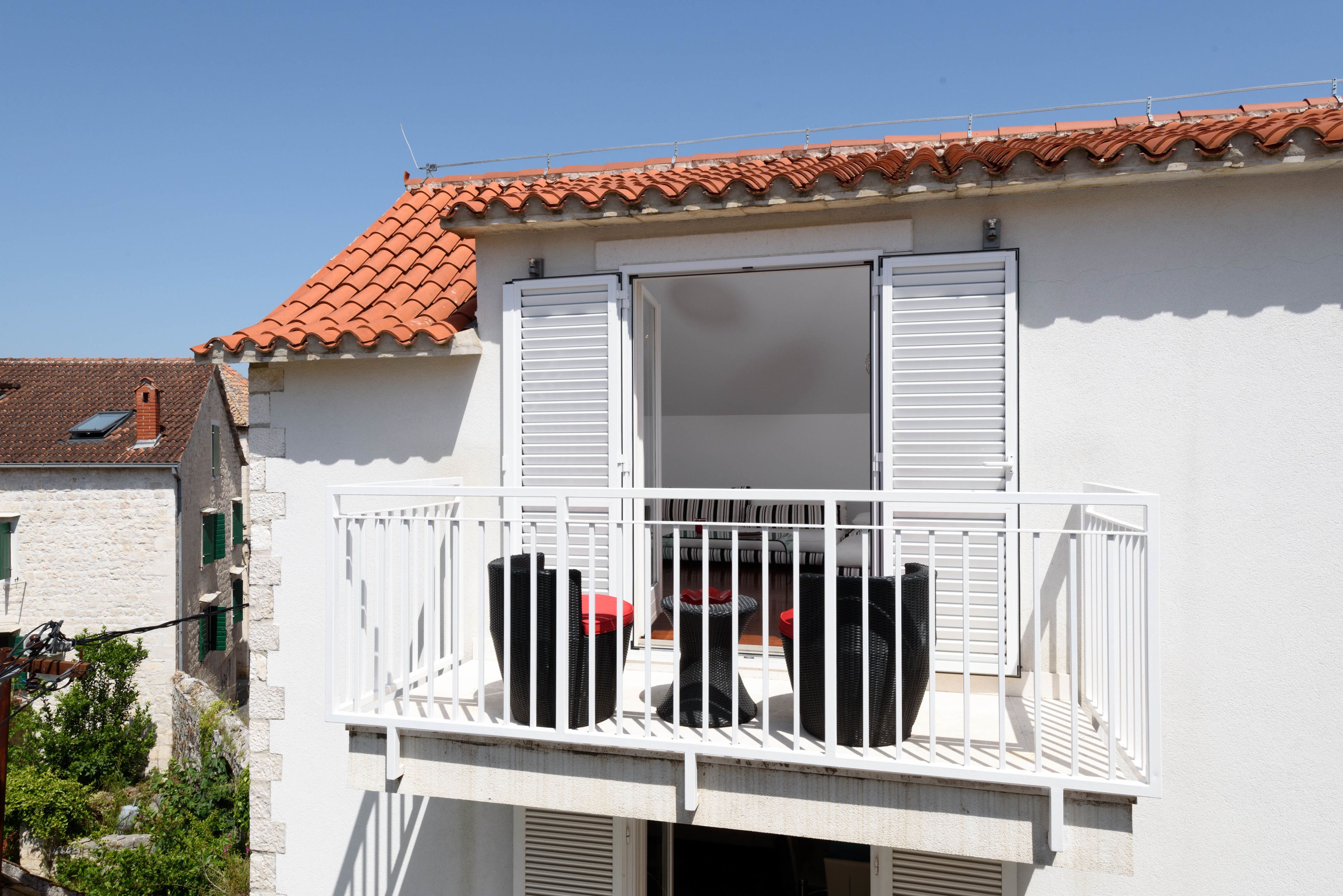 Un edificio blanco con techo de tejas rojas, un balcón con barandillas blancas y una ventana con contraventanas verdes.