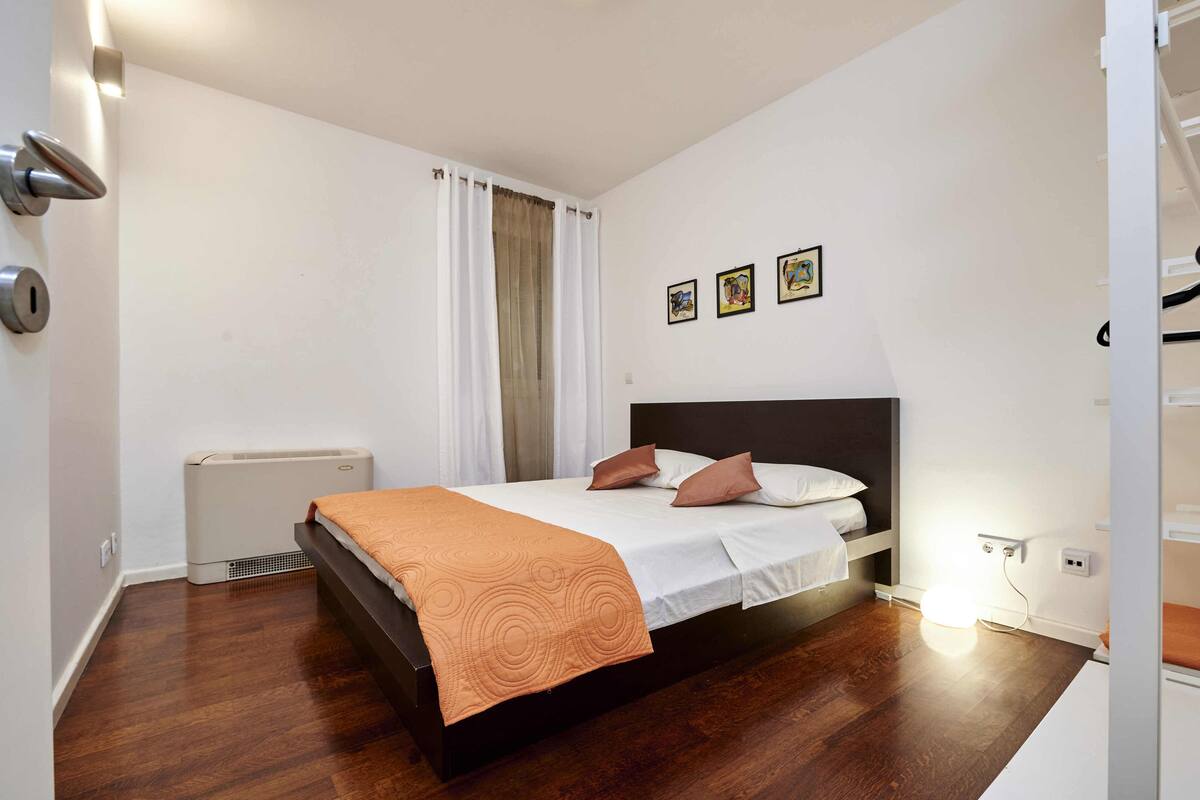 Quarto moderno com piso de madeira, cama com colcha branca e laranja, aquecedor e luminária fixada na parede.