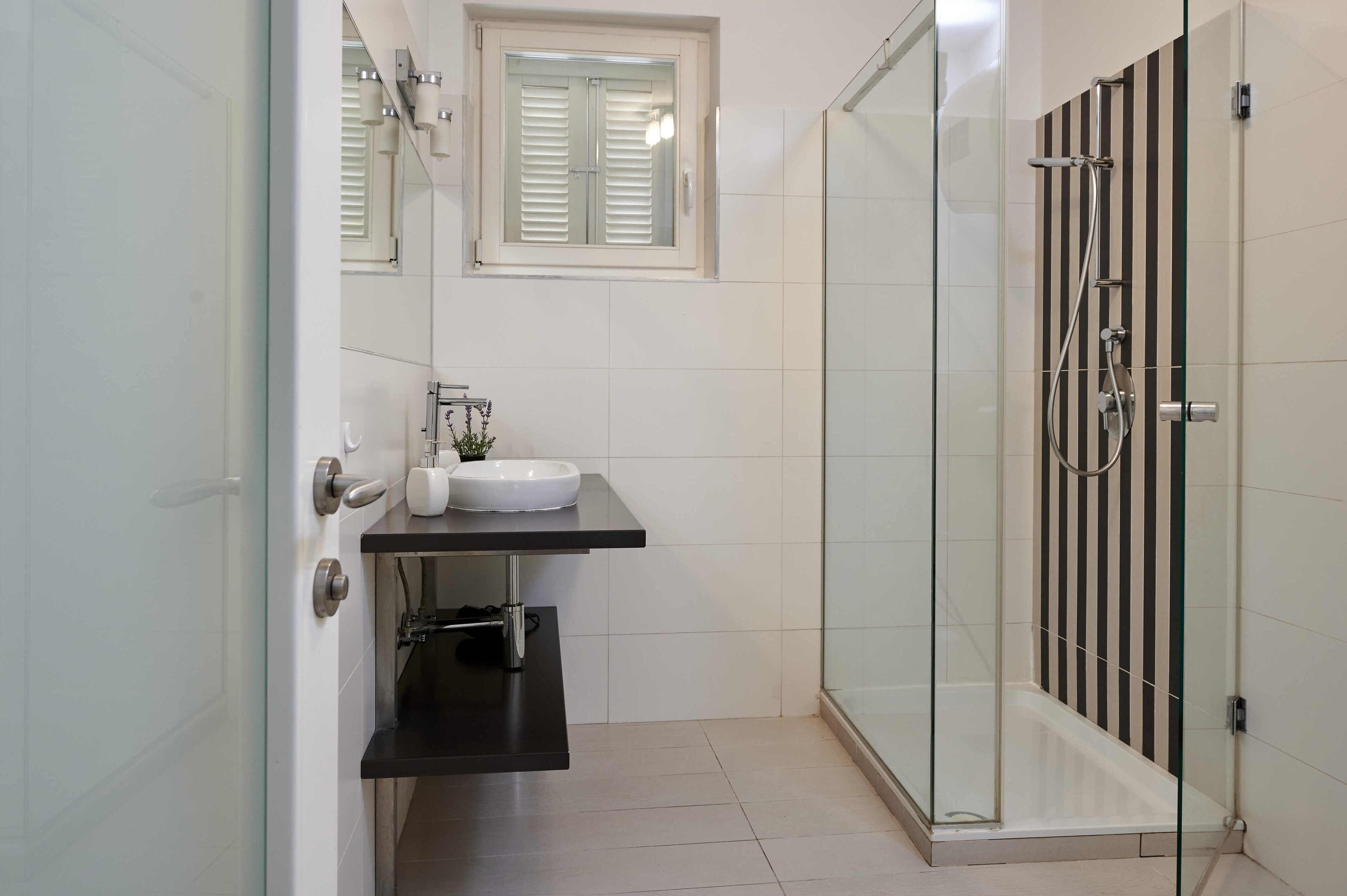 A modern bathroom with a glass shower enclosure, a white sink, and a window with blinds.