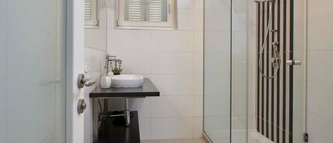 A modern bathroom with a glass shower enclosure, a white sink, and a window with blinds.