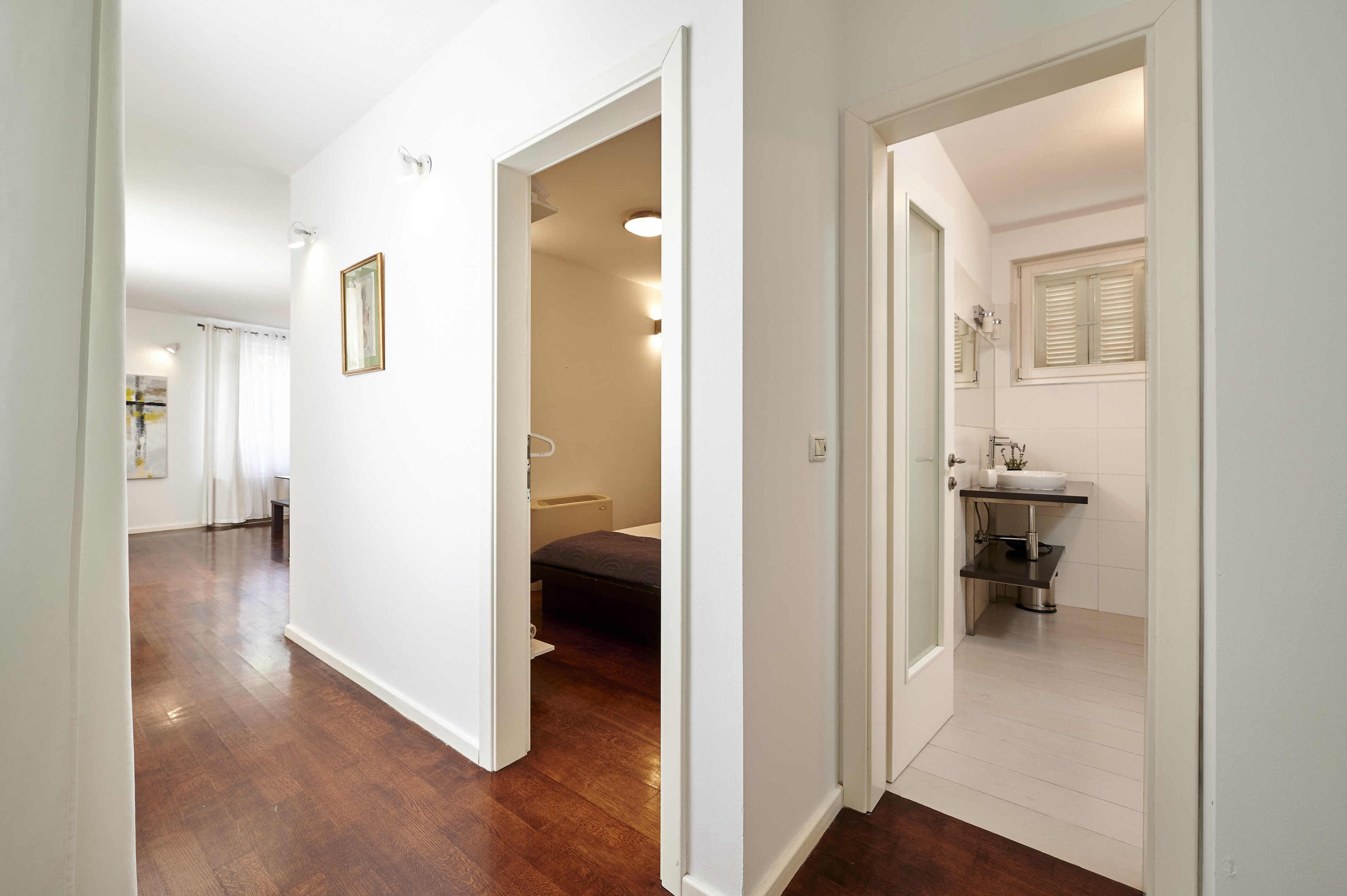 A hallway with a wooden floor leading to a bedroom with a bed and a bathroom with a sink and mirror.