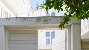 A narrow, enclosed outdoor corridor with a closed white shutter door, a small table with a plant, and a visible balcony in the background.