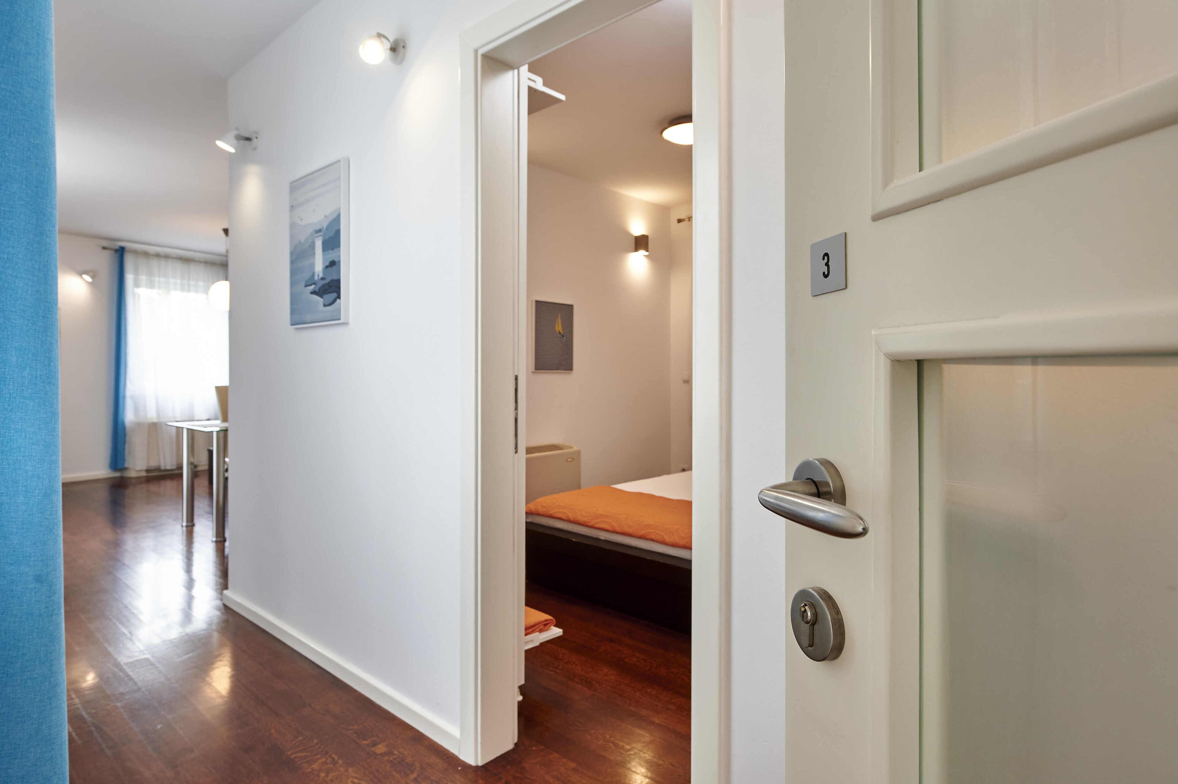 A hotel room with a bed, a small table, and a chair. There is a door with a number plate.