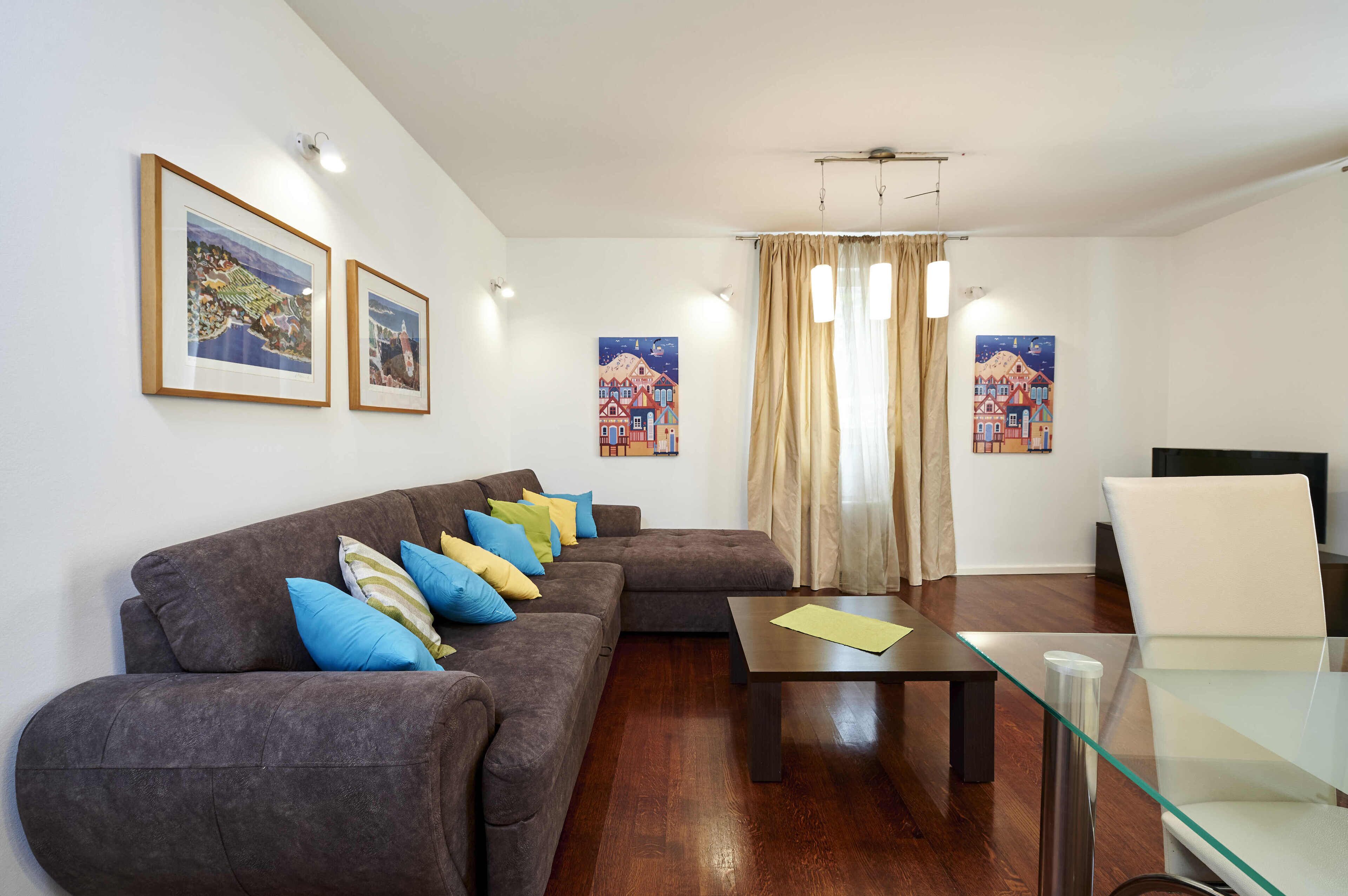 Una sala de estar con un sofá marrón, una mesa de centro de vidrio y una silla blanca. Hay cuadros enmarcados en la pared y una ventana con cortinas.