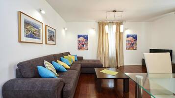 Una sala de estar con un sofá marrón, una mesa de centro de vidrio y una silla blanca. Hay cuadros enmarcados en la pared y una ventana con cortinas.