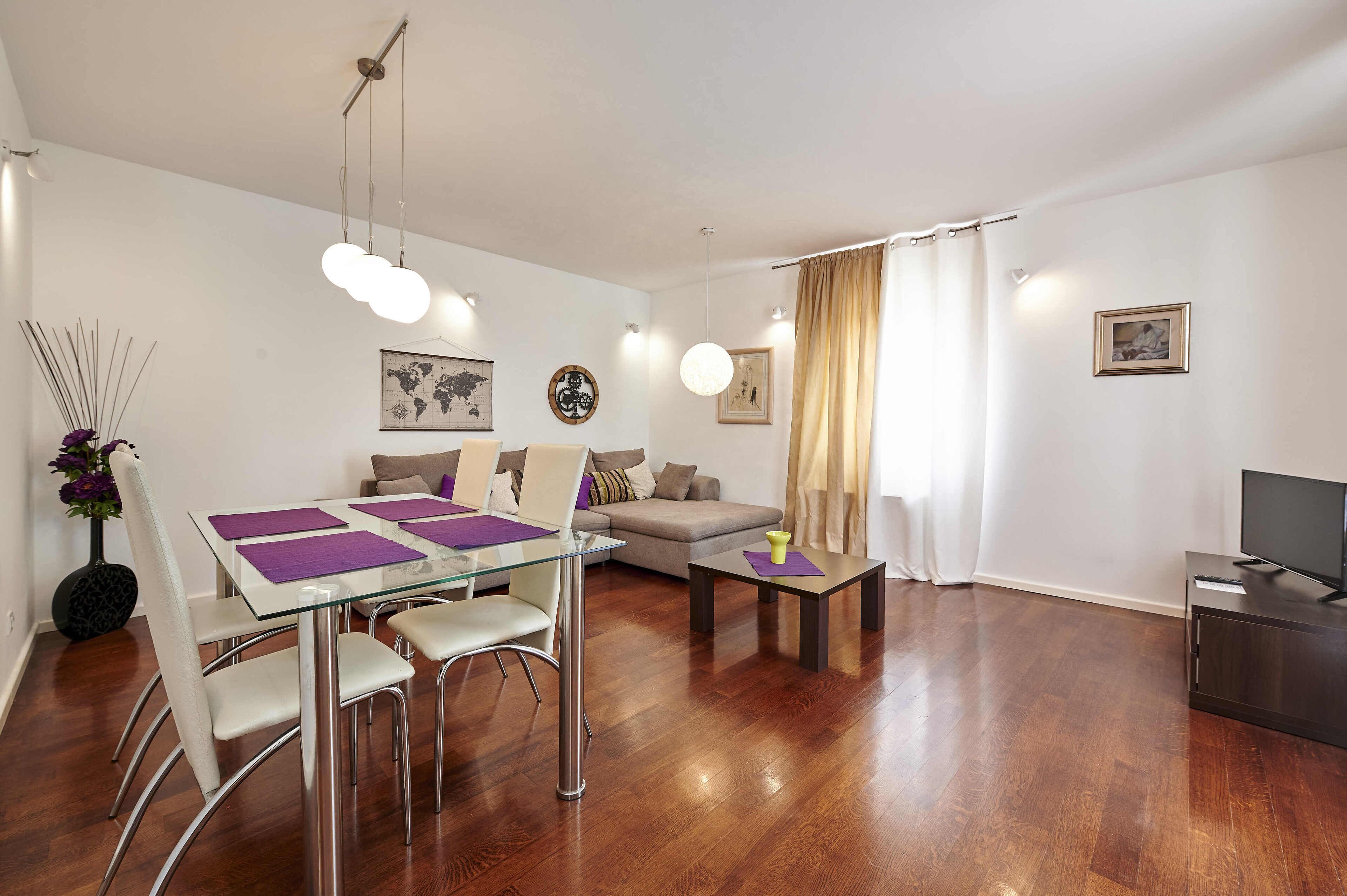 En moderne stue med et glasbord, hvide stole, en sofa, et sofabord og et tv-skab.