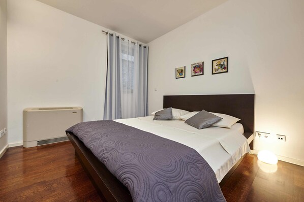 A bedroom with a bed, a radiator, a window with curtains, and three framed pictures on the wall.