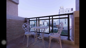 Ein Balkon mit Tisch und zwei Stühlen, Blick auf die Stadt durch große Fenster, und ein Gebäude im modernen Stil.