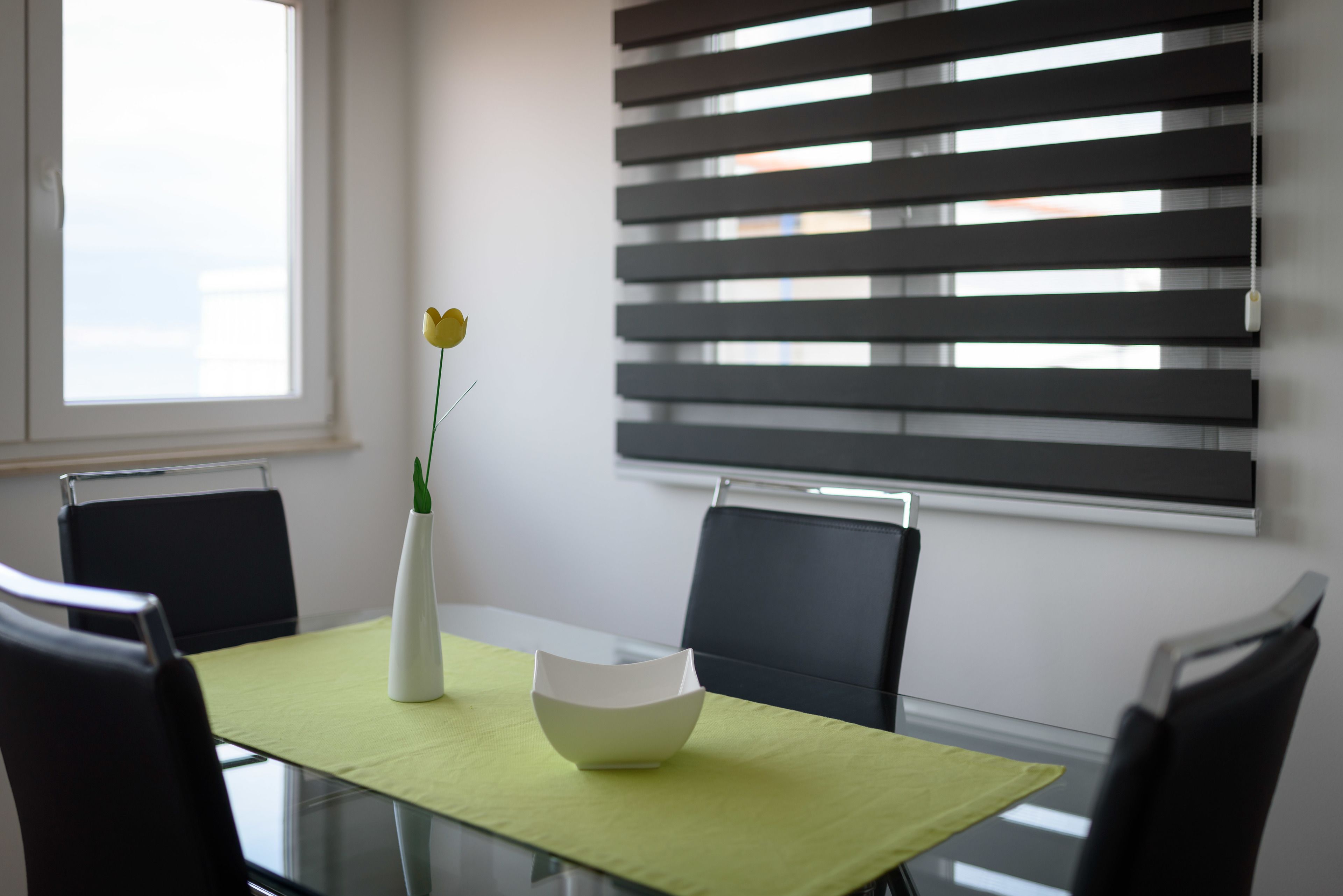 Un espace repas moderne avec une table en verre, des chaises noires, un vase blanc contenant une fleur jaune et un bol blanc.