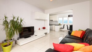 Een moderne woonkamer met een flatscreen televisie, een zwarte leren bank met kleurrijke kussens, een witte tv-meubel en een groene plant in pot.