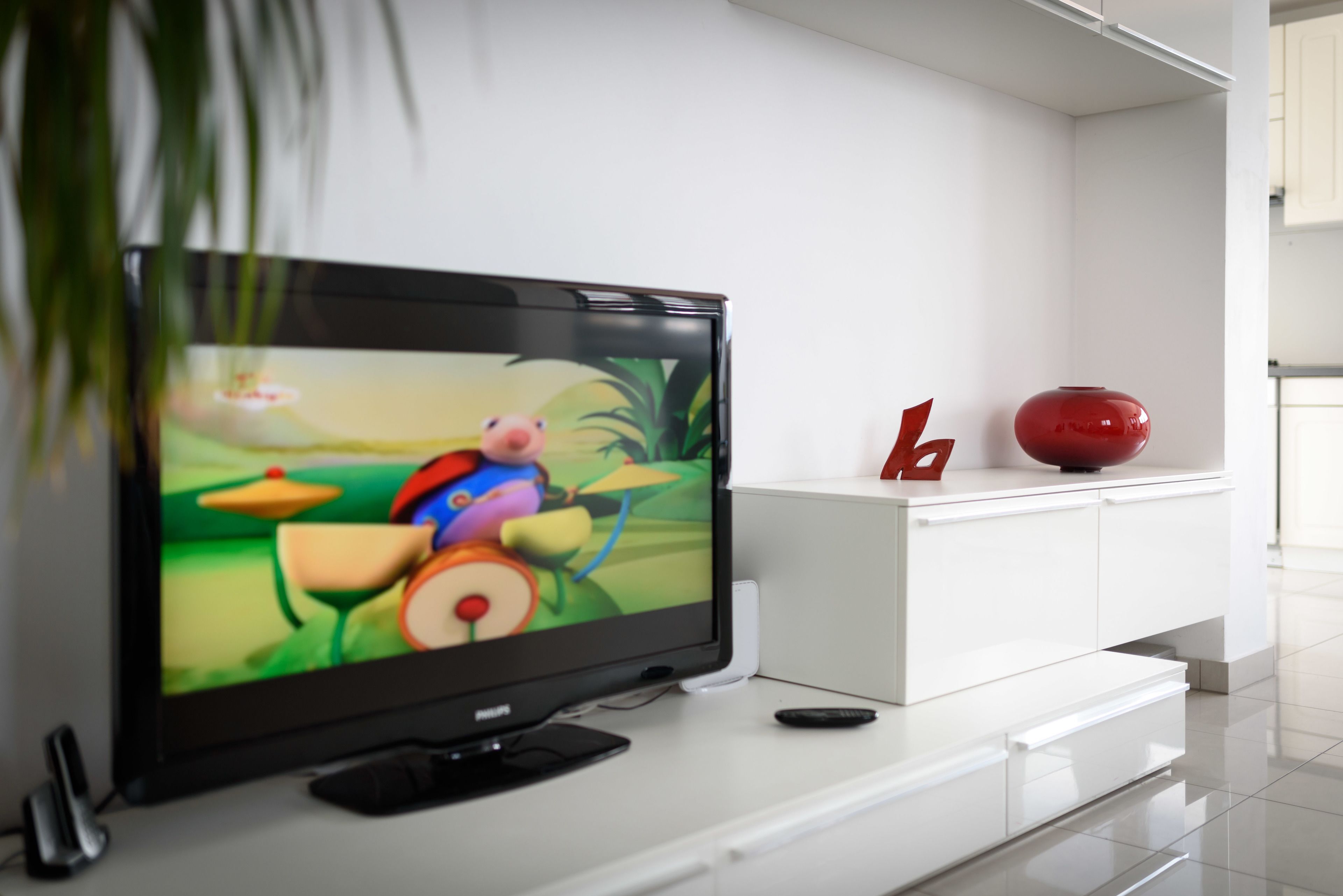 A flat-screen TV on a white entertainment unit with a remote control, a decorative red vase, and a small plant.