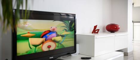 A flat-screen TV on a white entertainment unit with a remote control, a decorative red vase, and a small plant.