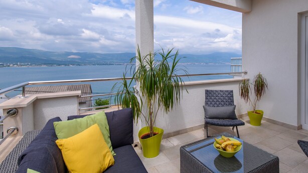 Een balkon met een bank, stoelen en een tafel met fruit. Er staan potplanten en er is uitzicht op zee en bergen.