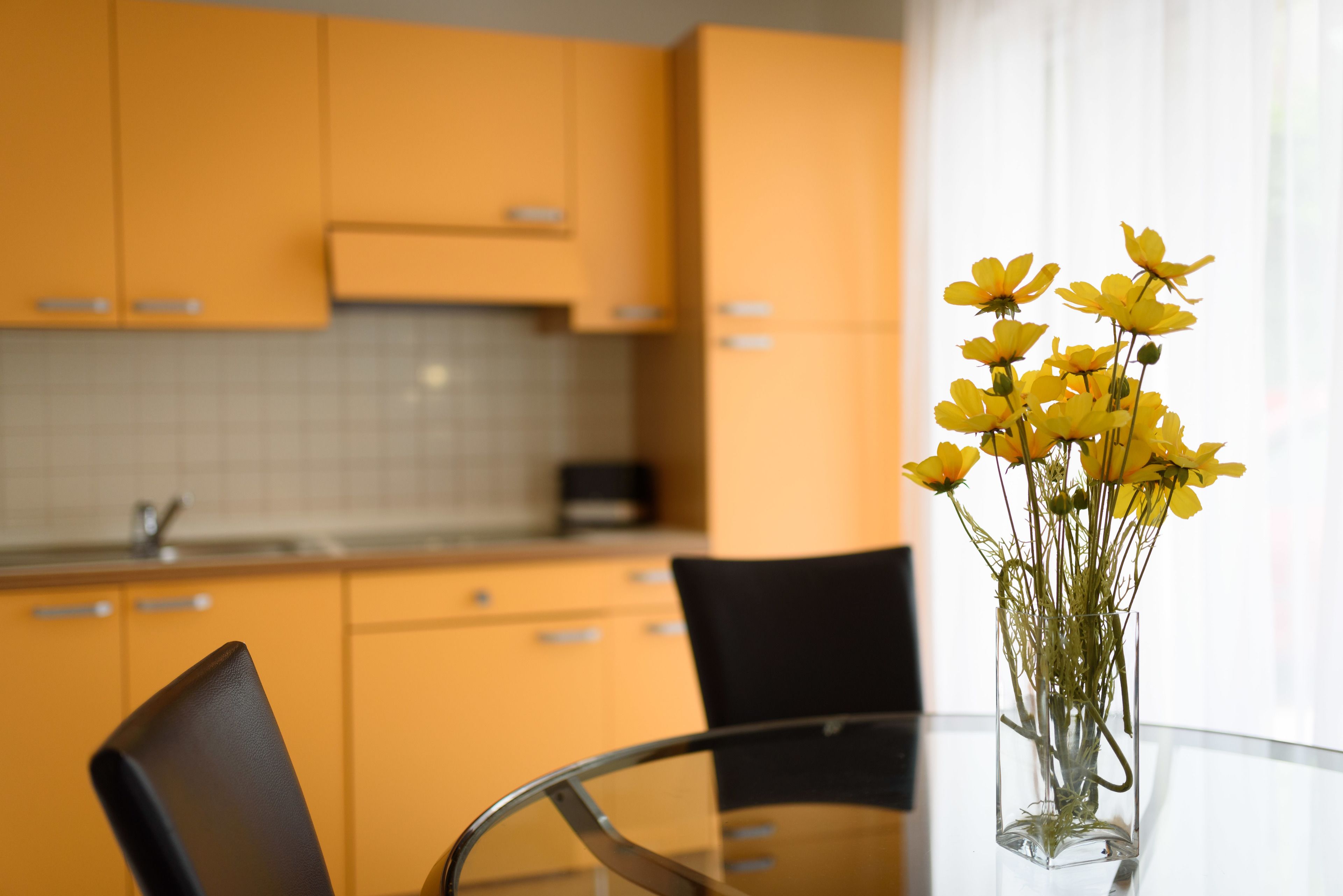 A modern kitchen with yellow cabinets, a glass table, and a vase with yellow flowers.