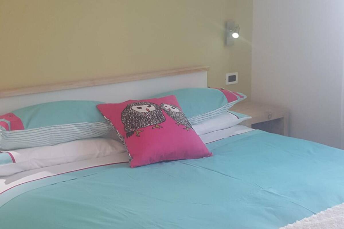 A neatly made bed with a teal bedspread, a pink owl pillow, and white pillows. A colorful painting of owls and flowers hangs on the wall above the bed.