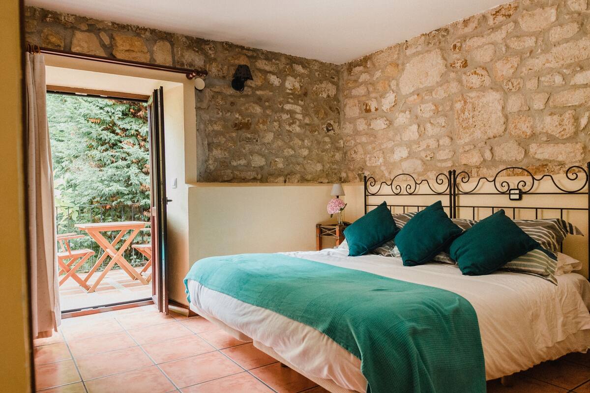 A bedroom with a stone wall, a bed with green and white bedding, a nightstand with a lamp, and a view of a garden through an open door.