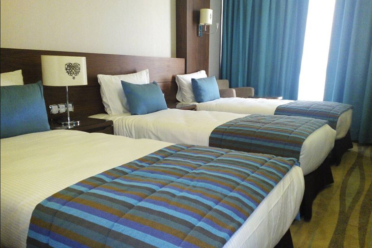 Hotellihuone, jossa on kolme sänkyä. Jokaisessa sängyssä on raidallinen peitto, yöpöydällä on lamppu ja sängyn päädyssä on koristepaneeli.