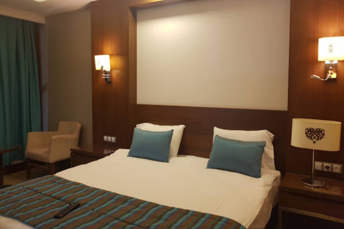 Hotellihuone, jossa on suuri sänky, kaksi tyynyä, raidallinen peitto, tuoli, yöpöytä ja seinään kiinnitetyt lamput.