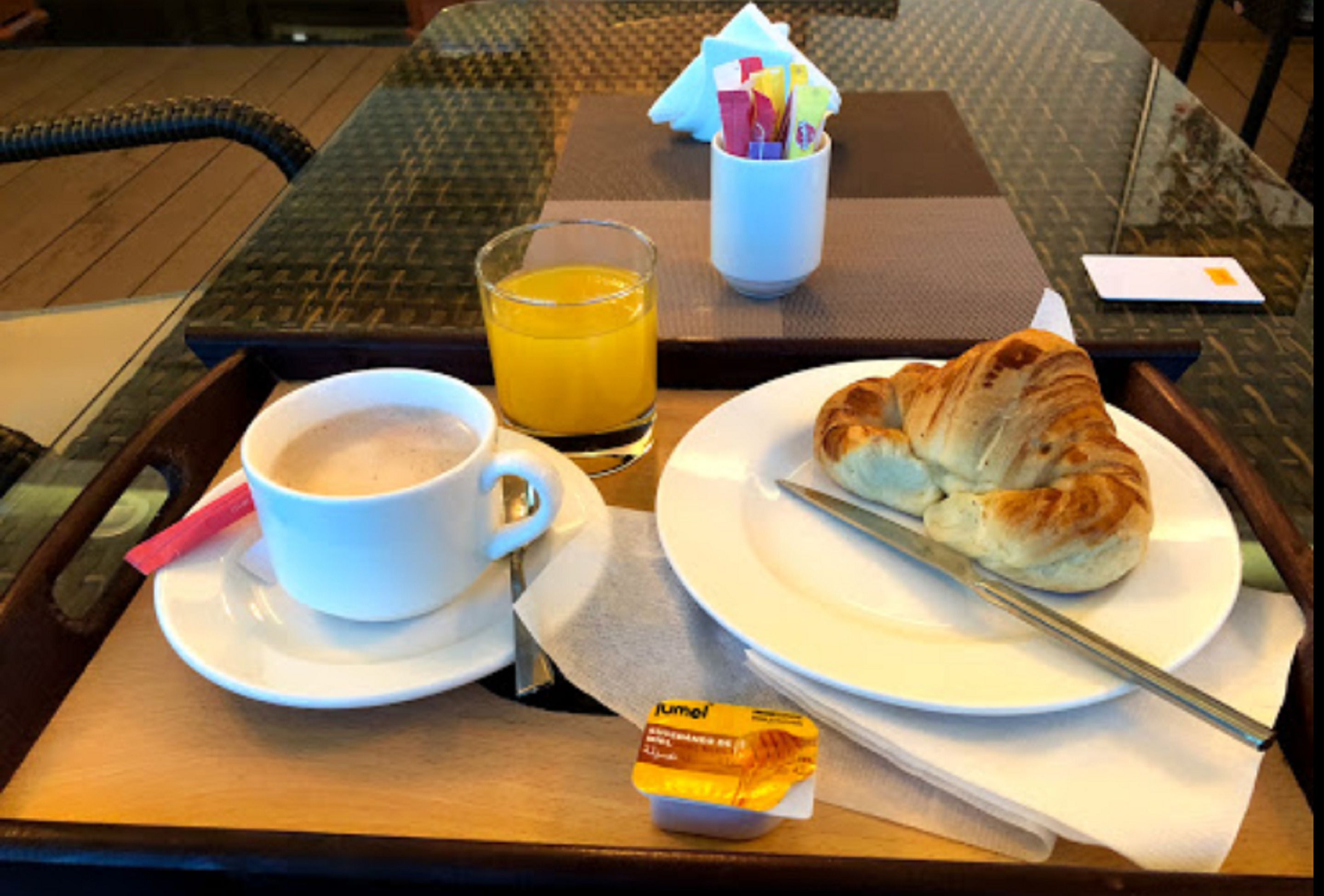Una bandeja de desayuno con un croissant, una taza de café y un vaso de jugo de naranja sobre una mesa de madera.