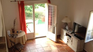 A room with a tiled floor, a dining table with chairs, a TV, a lamp, and a sliding glass door leading to an outdoor area. - Riad des Vignes (Ramatuelle)