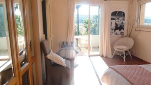 A bedroom with a bed, a chair, a small table, and a view of the outdoors. - Riad des Vignes (Ramatuelle)