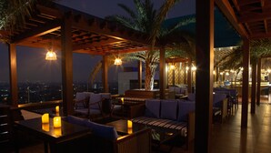Een terras met tafels en stoelen, een pergola en een uitzicht op de stad in de avond.