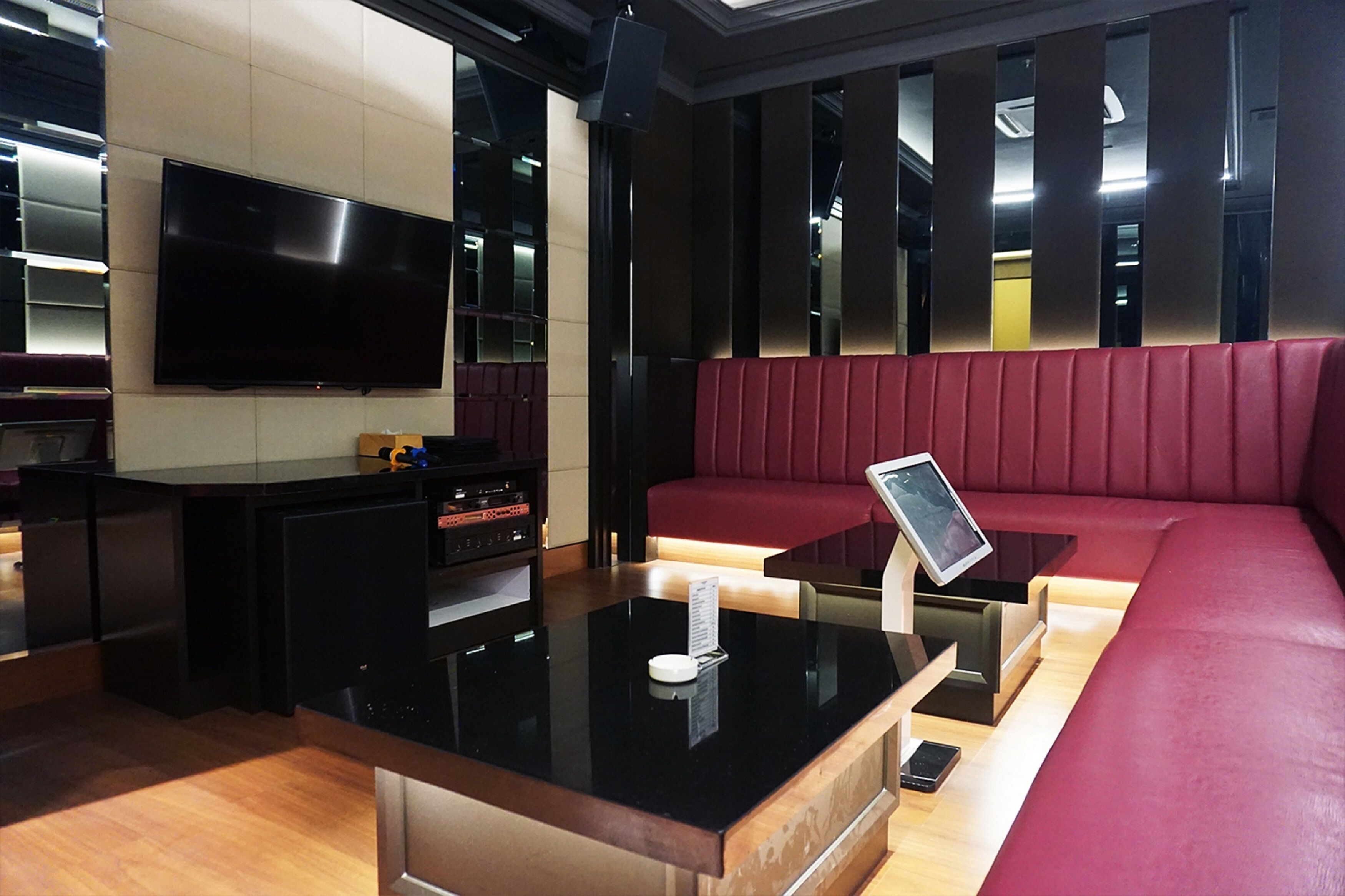 Un interior moderno con un mostrador de recepción negro, un televisor de pantalla plana, una zona de estar con cojines rojos y un suelo de madera.