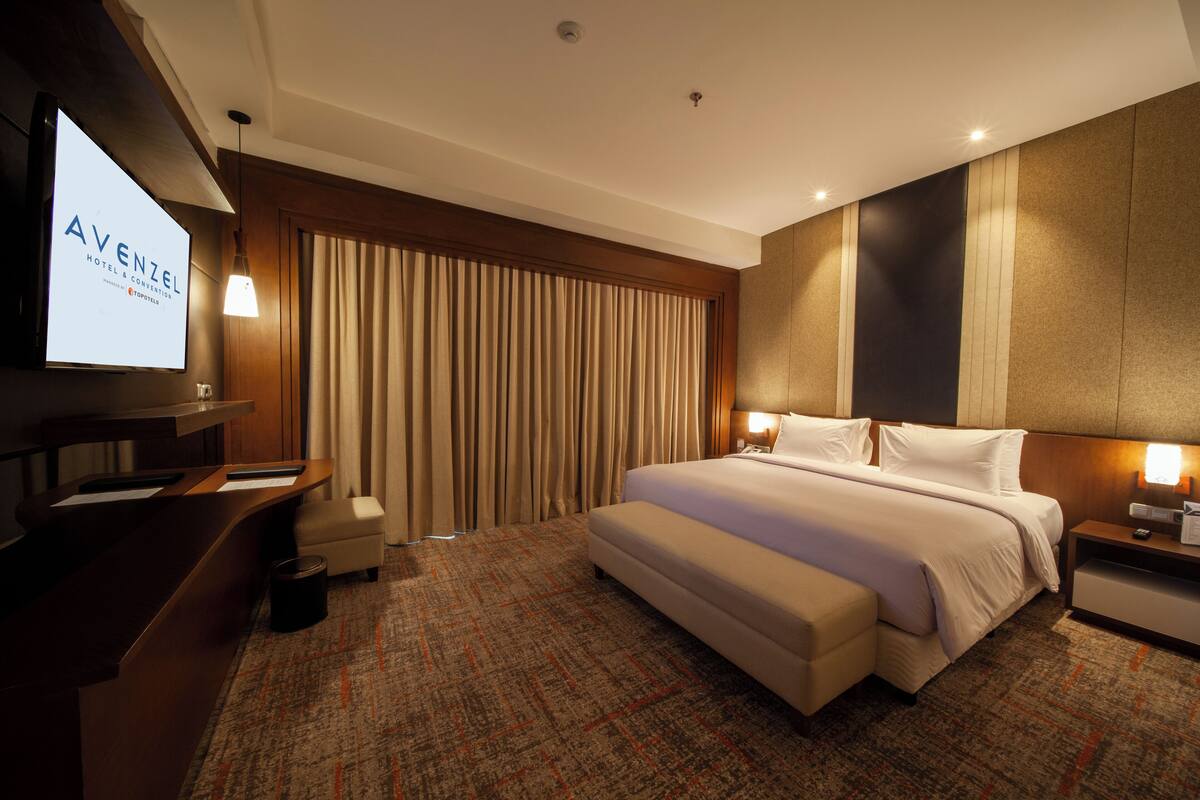 Ein Hotelzimmer mit einem großen Bett, einem Schreibtisch, einem Fernseher und Blick auf Vorhänge.