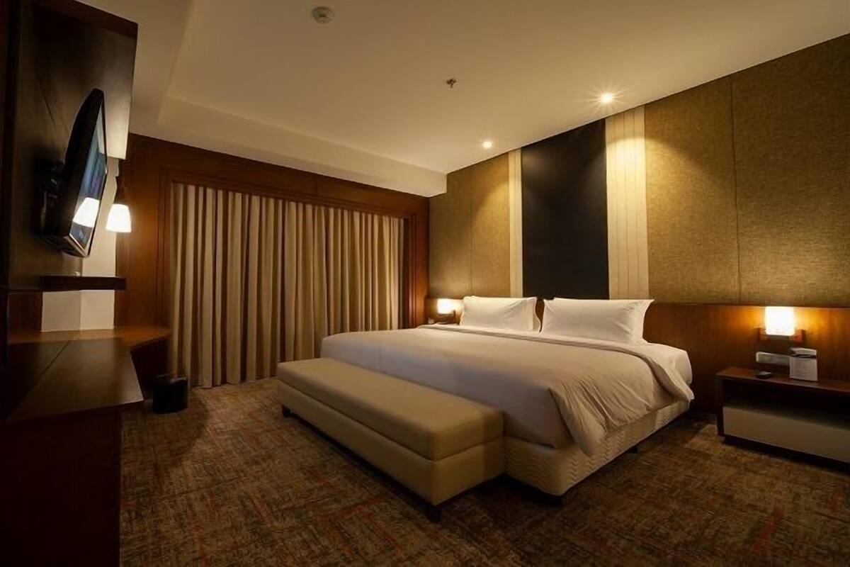 Ein Hotelzimmer mit einem großen Bett, einem flachbildfernseher an der Wand, einem Nachttisch mit Lampe und einem großen Fenster mit Vorhängen.