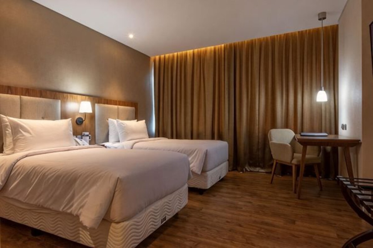 Une chambre d’hôtel avec deux lits, un bureau, une chaise et une grande fenêtre avec des rideaux.