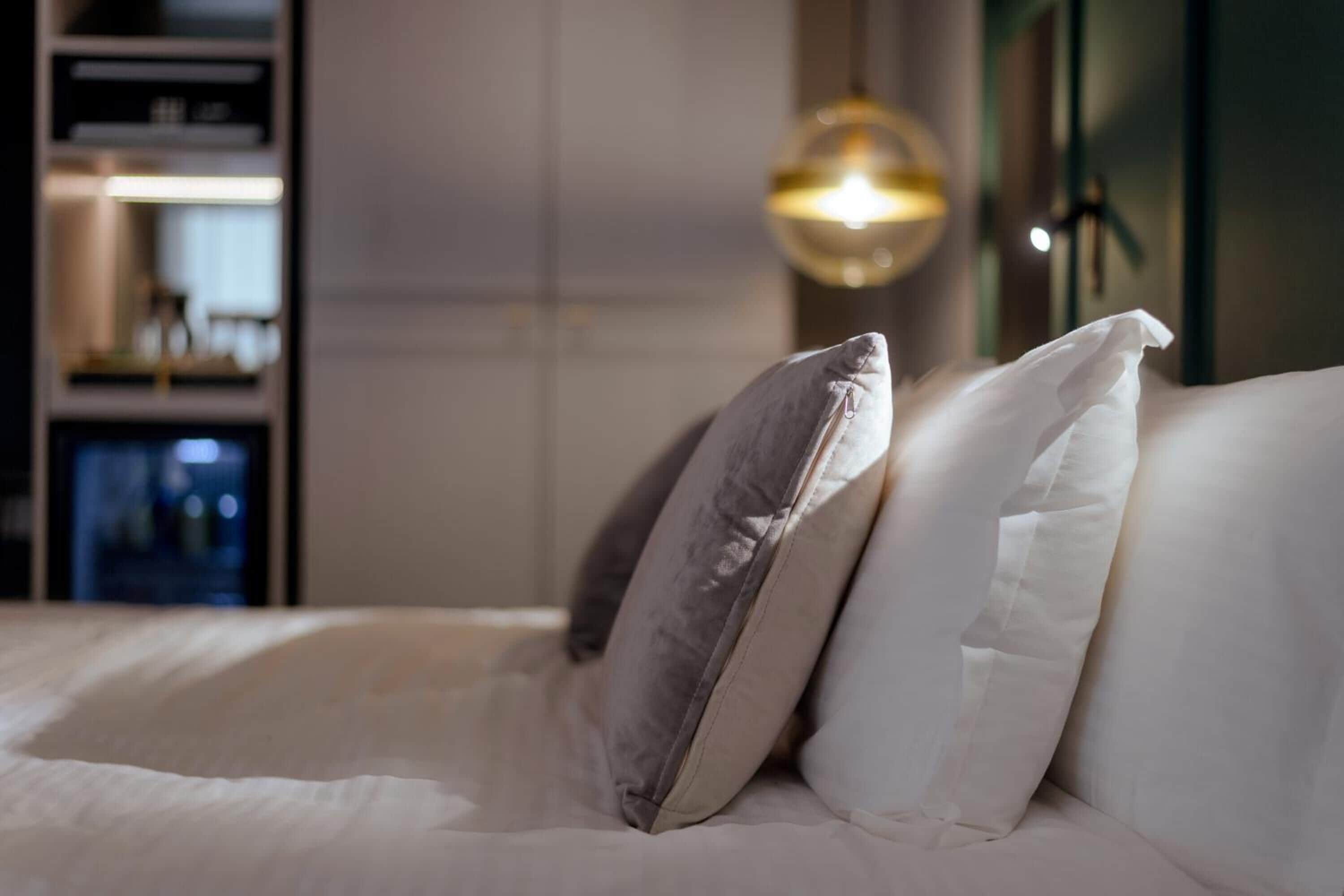 A neatly made bed with white and gray pillows, a modern lamp, and a shelf with items in the background.