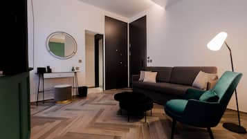 A modern living room with a herringbone wood floor, a dark green armchair, a black coffee table, a grey sofa, and a round mirror on the wall.