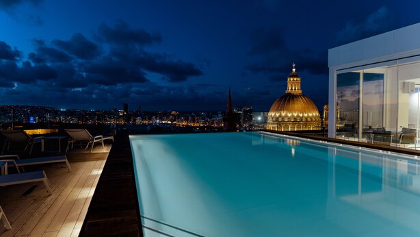 Una piscina en la azotea con sillones de descanso, un edificio moderno con grandes ventanales y un horizonte urbano con una cúpula destacada al fondo.