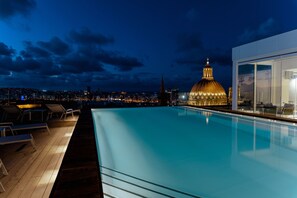 Una piscina en la azotea con sillones de descanso, un edificio moderno con grandes ventanales y un horizonte urbano con una cúpula destacada al fondo.