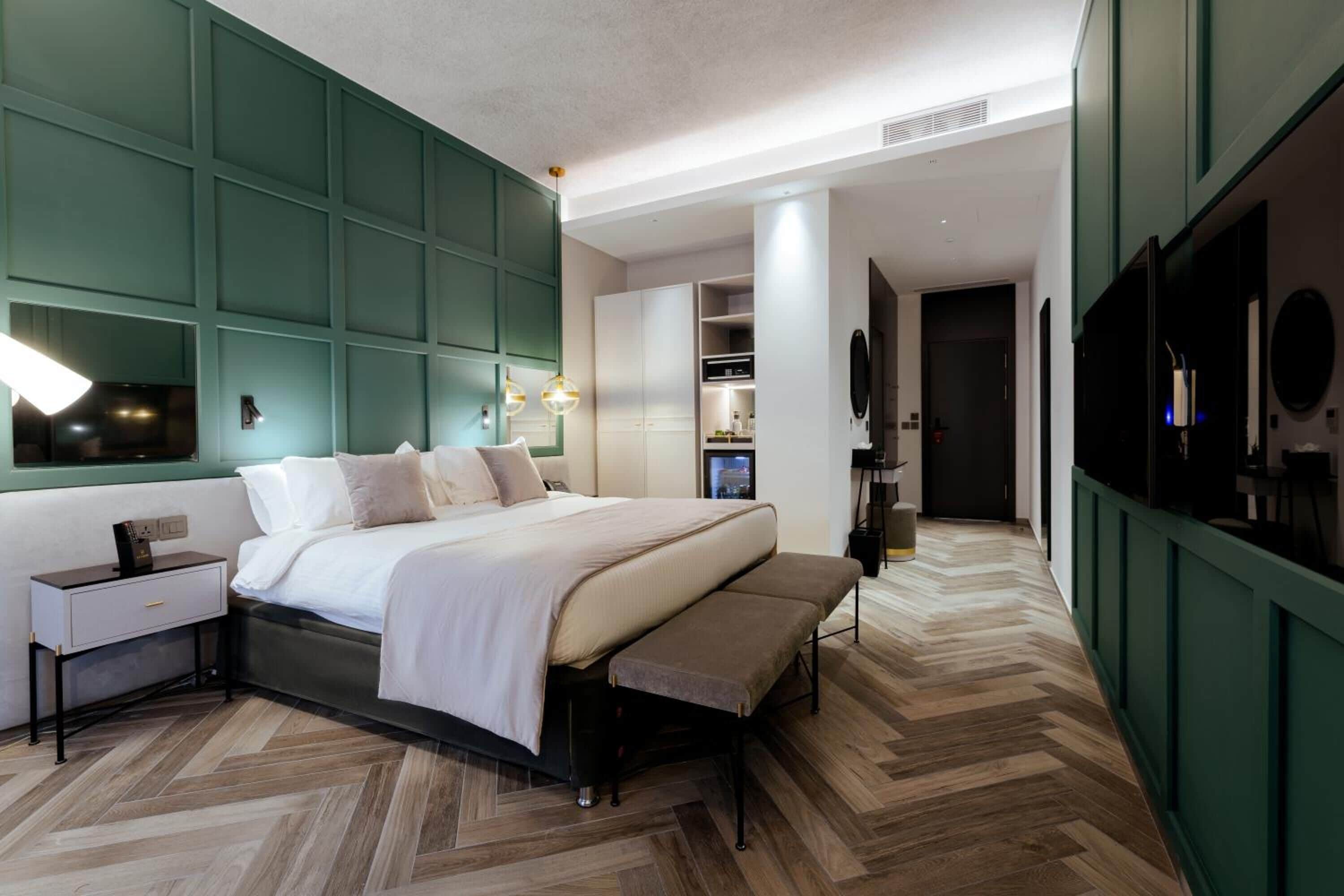 A modern bedroom with a large bed, bedside tables, a bench, and built-in green storage.