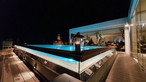 Una piscina con borde de cristal, una terraza de madera y un edificio con fachada de vidrio al fondo.