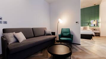 A modern room with a dark brown sofa, a round wooden table, and a teal armchair. A floor lamp is on, and a bed with a green headboard is visible in the background.