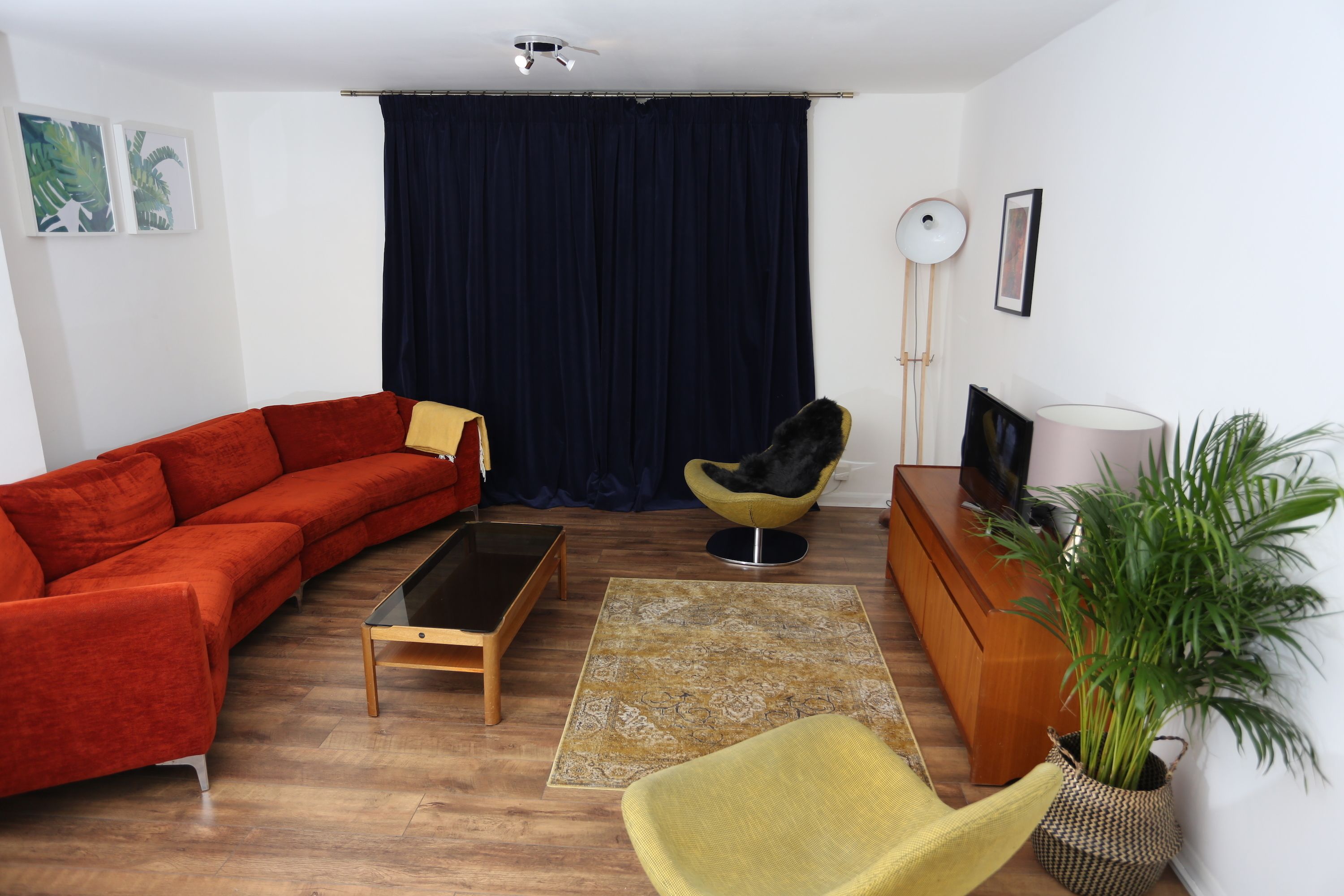 A living room with a red sofa, a yellow armchair, a wooden coffee table, a floor lamp, a TV stand, and a potted plant.
