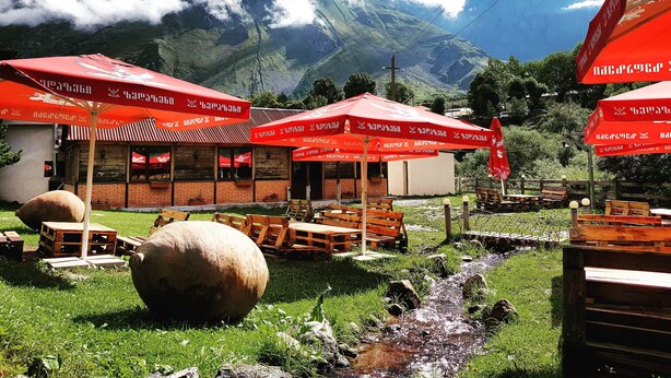A restaurant with outdoor seating, red umbrellas, and a mountain backdrop. - Cozy Hotel Kazbegi (Kazbegi)