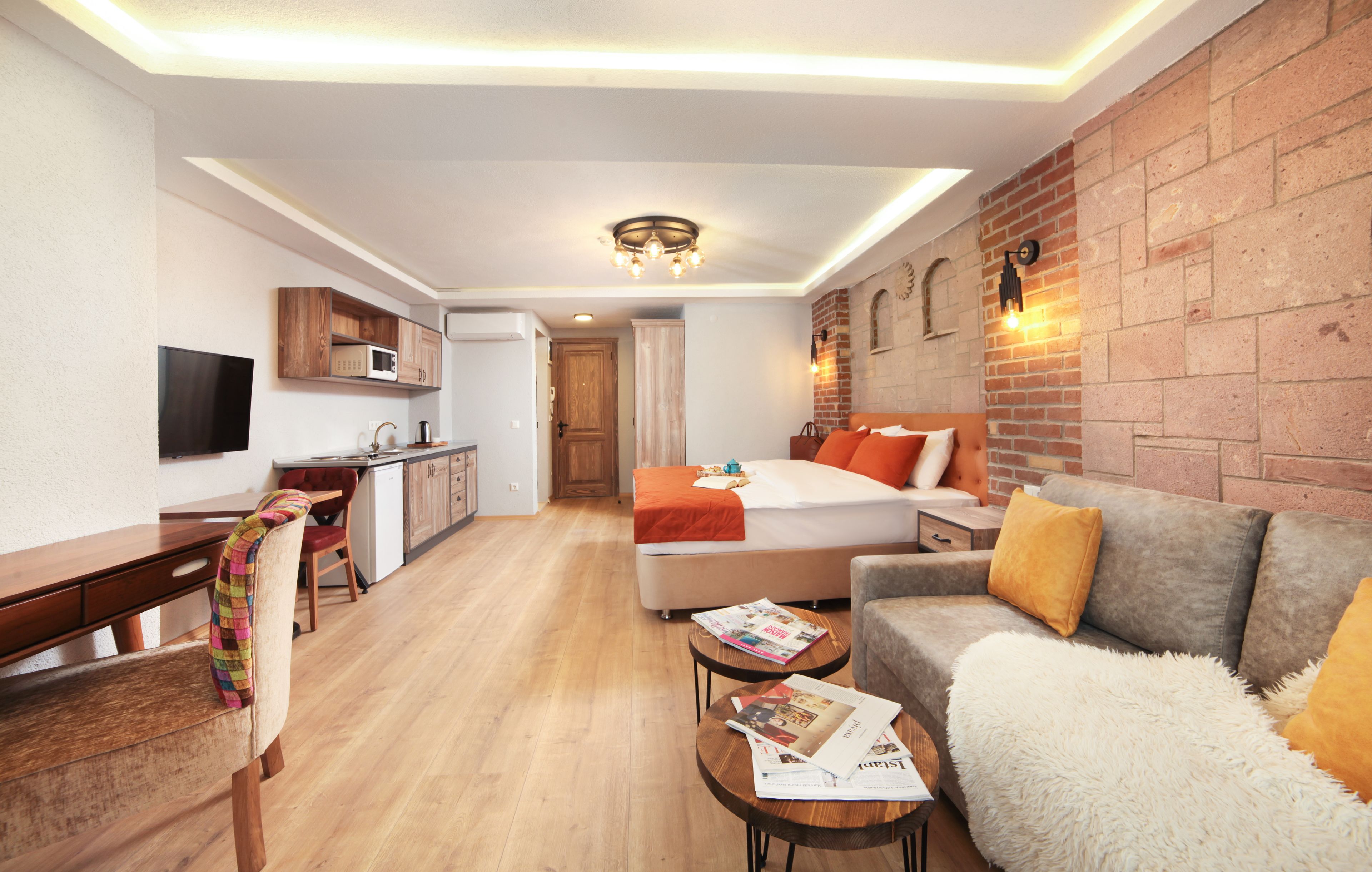 Una habitación de hotel compacta con kitchenette, una cama con ropa de cama naranja, un sofá gris con una manta blanca esponjosa y una mesa de comedor de madera con sillas.