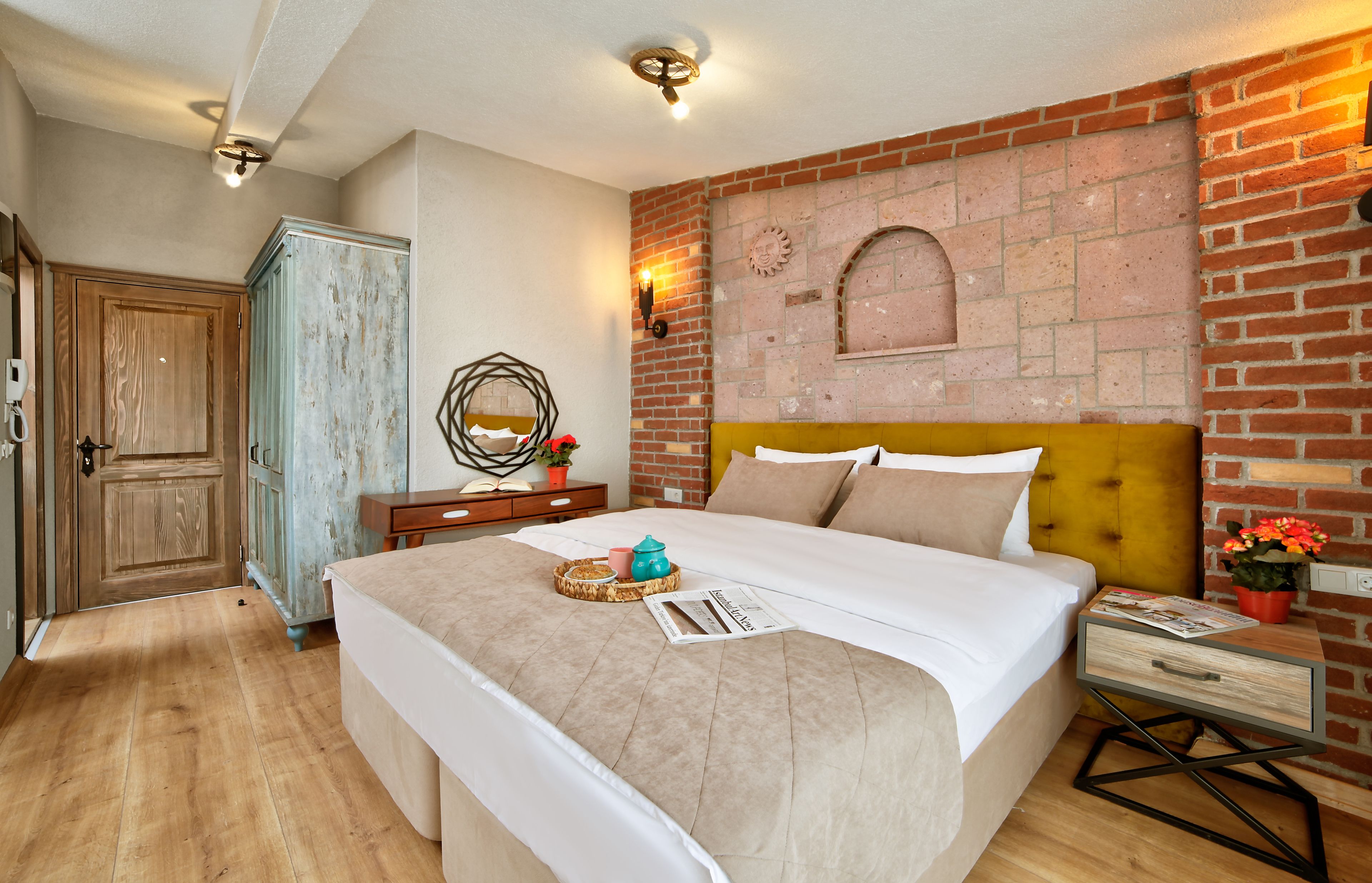 A bedroom with a brick wall, a wooden headboard, a wooden wardrobe, a bedside table with a magazine, and a small potted plant.