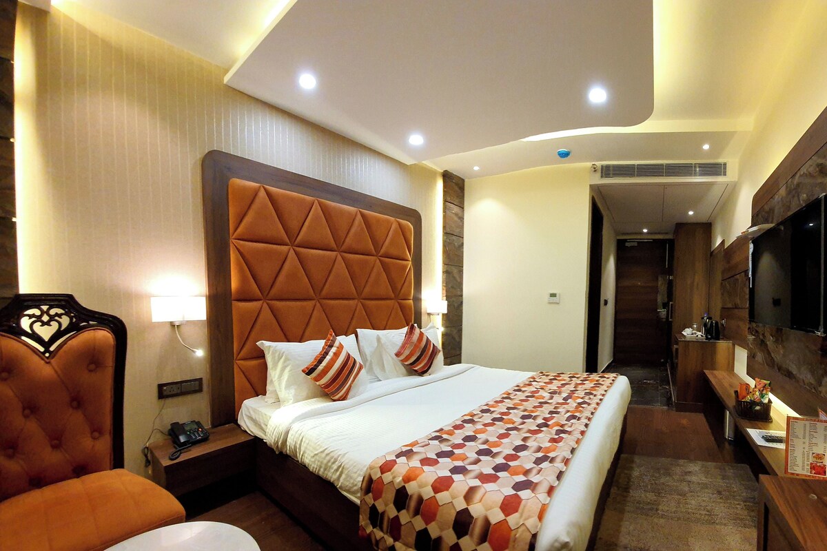 Habitación de hotel con una cama grande, cabecera de madera, mesita de noche con lámpara, televisor montado en la pared y un pequeño escritorio con un menú.