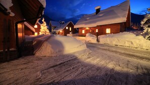 Vila coberta de neve com casas de madeira, uma árvore de Natal iluminada e céu claro ao entardecer.