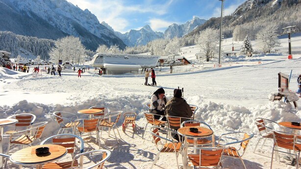 Uma área externa com mesas e cadeiras coberta de neve, um teleférico ao fundo e montanhas à distância.