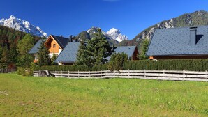 Paisagem rural com casas de madeira, cerca de madeira e montanhas ao fundo.
